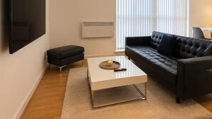 A modern living room with a flat-screen TV, a black leather sofa, a white coffee table, and a large window with blinds. - Webster Road Park Loft Apartment (Liverpool)