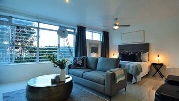 A modern living room with a sofa, a coffee table, a bed, and a view of the cityscape through large windows.
