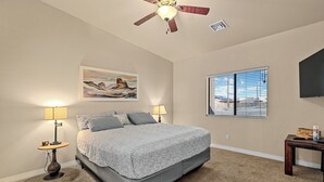 A bedroom with a bed, two bedside lamps, a ceiling fan, a painting, a window with blinds, and a television on the wall. - Sabino Gorgeous Heatable Pool Retreat (Lake Havasu City)