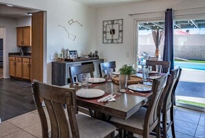 Uma sala de jantar com uma mesa posta para quatro pessoas, cadeiras de madeira, um relógio na parede e vista para o exterior.