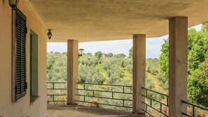 A covered outdoor area with concrete pillars, a railing, and a view of olive trees and a distant building.