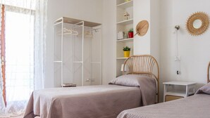 A bedroom with two beds, a wardrobe, shelves with plants, and a bedside table.