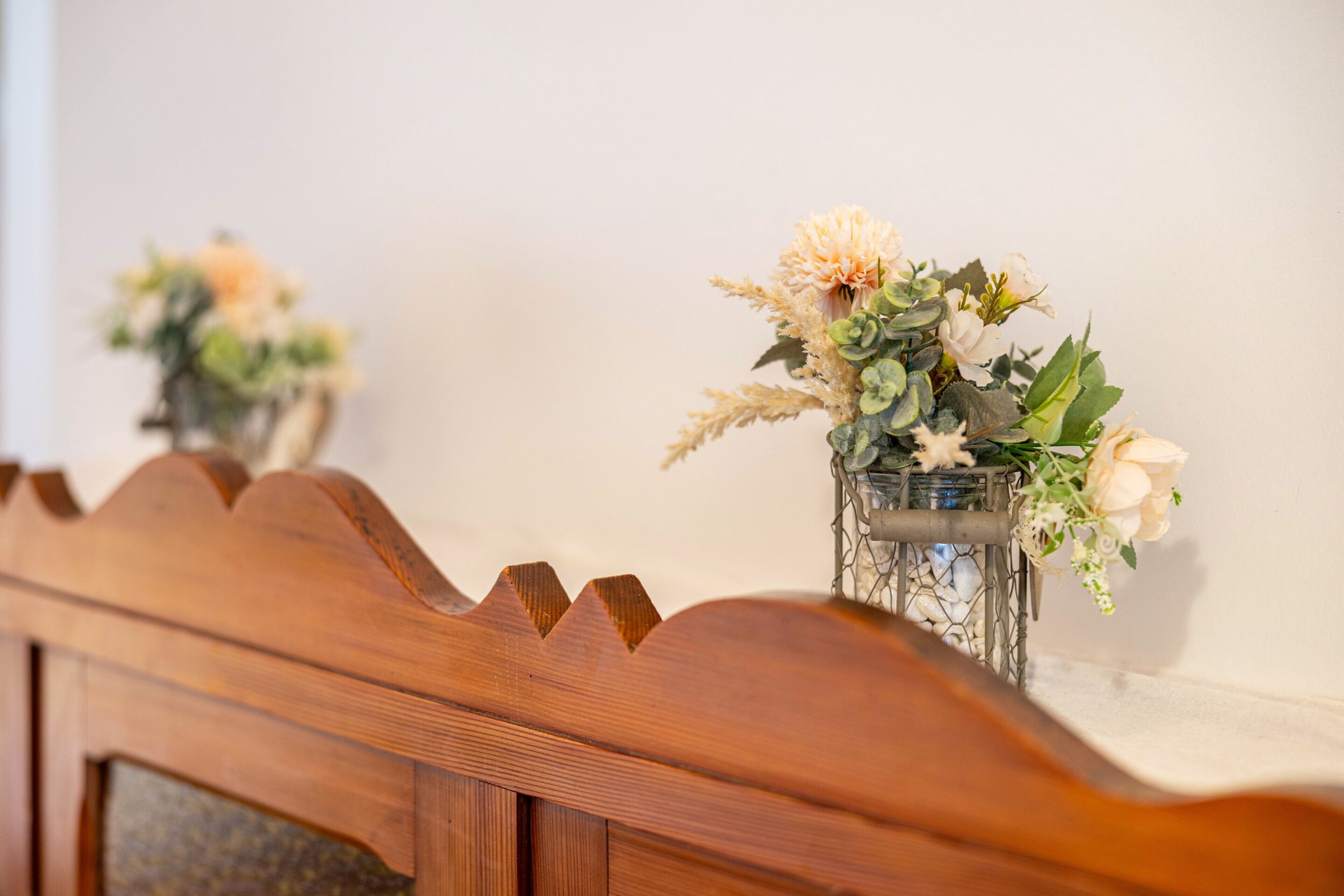 Un banco de madera con un respaldo decorativo, un espejo fijado a la pared y un jarrón con flores.
