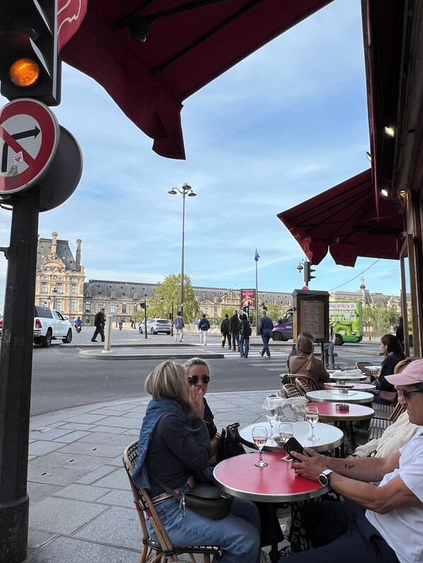 Terrasse mit Tischen und Stühlen, Straßenansicht mit Fußgängern und ein Gebäude im Hintergrund mit klassischer Architektur.