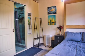 A bedroom with a bed, a radiator, a black nightstand, a wall-mounted shelf, and a door leading to a shower area. - Cozy Loft studio with views of Downtown Tijuana (Tijuana)
