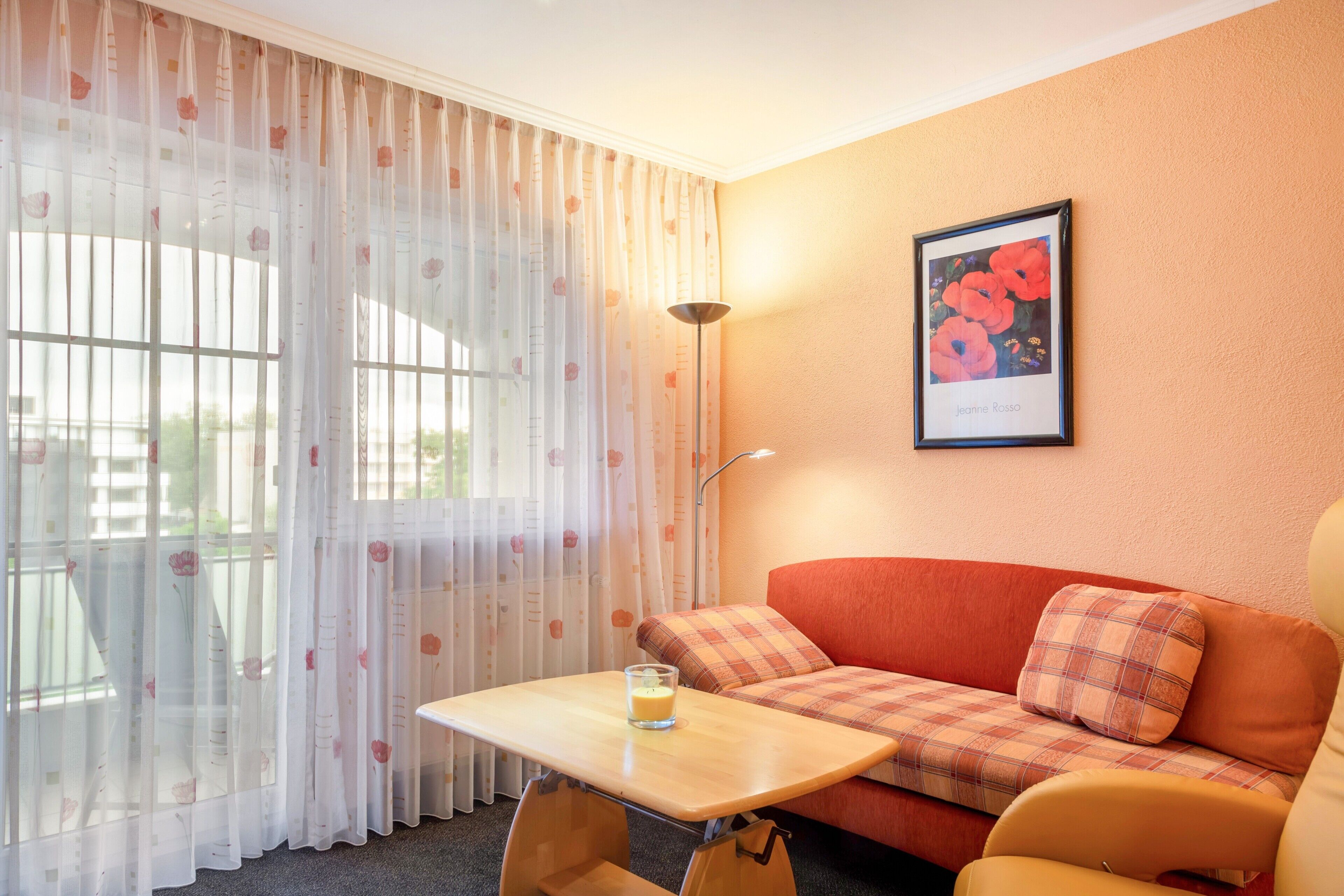 A room with a window, a table with a glass of water, a sofa, and a framed picture on the wall.