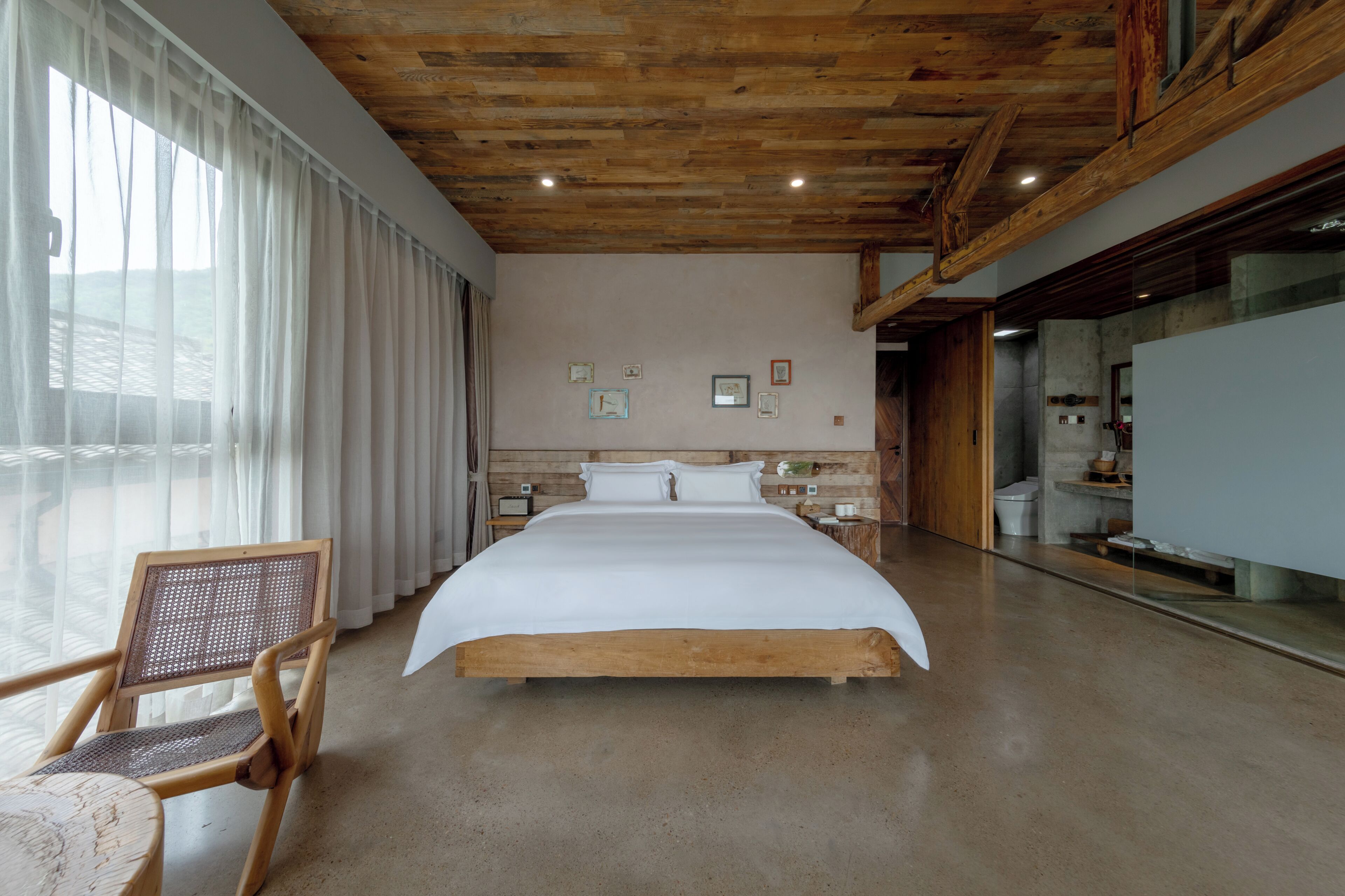 Een slaapkamer met een groot bed, houten plafond en een stoel bij het raam.