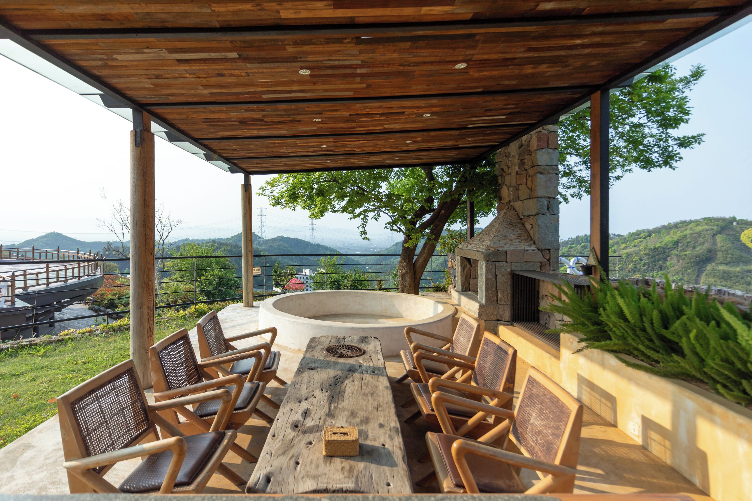 An outdoor patio with a wooden table and chairs, a stone fire pit, and a view of the surrounding landscape.