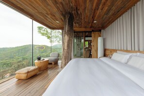 A bedroom with a large bed, wooden flooring, a glass wall with a view of green hills, and a wooden ceiling.