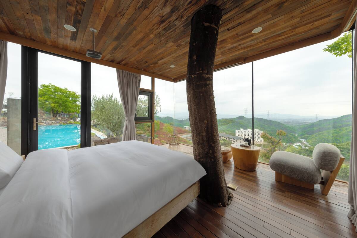 A bedroom with a large bed, a wooden ceiling, a view of a swimming pool, and a large tree trunk.