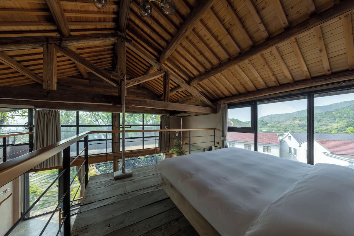 A bedroom with a wooden ceiling, a large bed, a balcony with a railing, and a view of buildings and greenery outside.