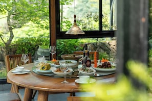A dining table set with various dishes, glasses, and a bottle, with a view of greenery outside.