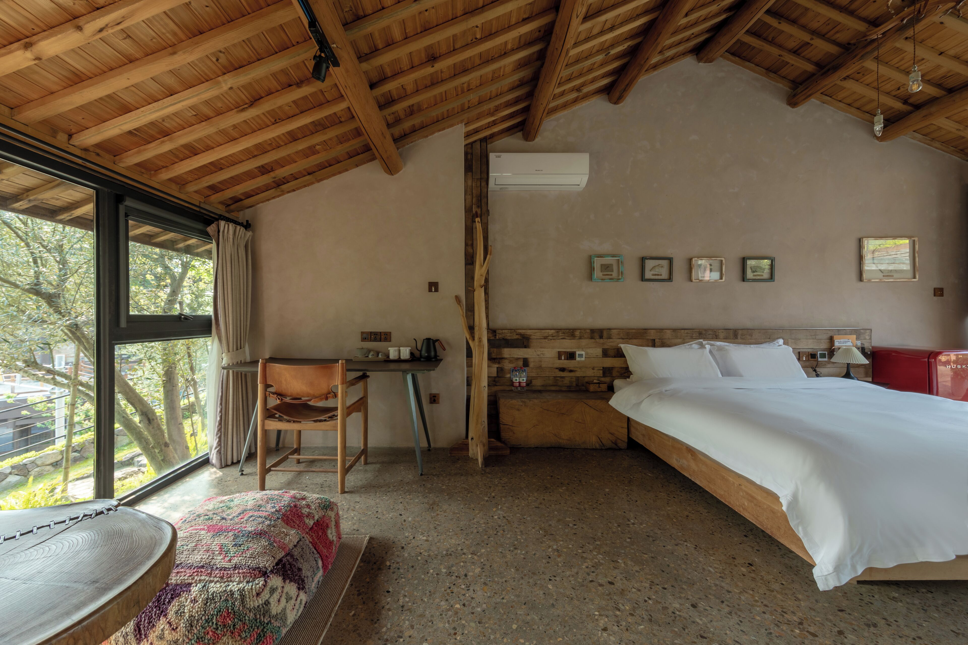 Ein Schlafzimmer mit Holzboden, einem Bett, einem Schreibtisch, einem Stuhl und einem Fenster mit Blick auf Bäume.