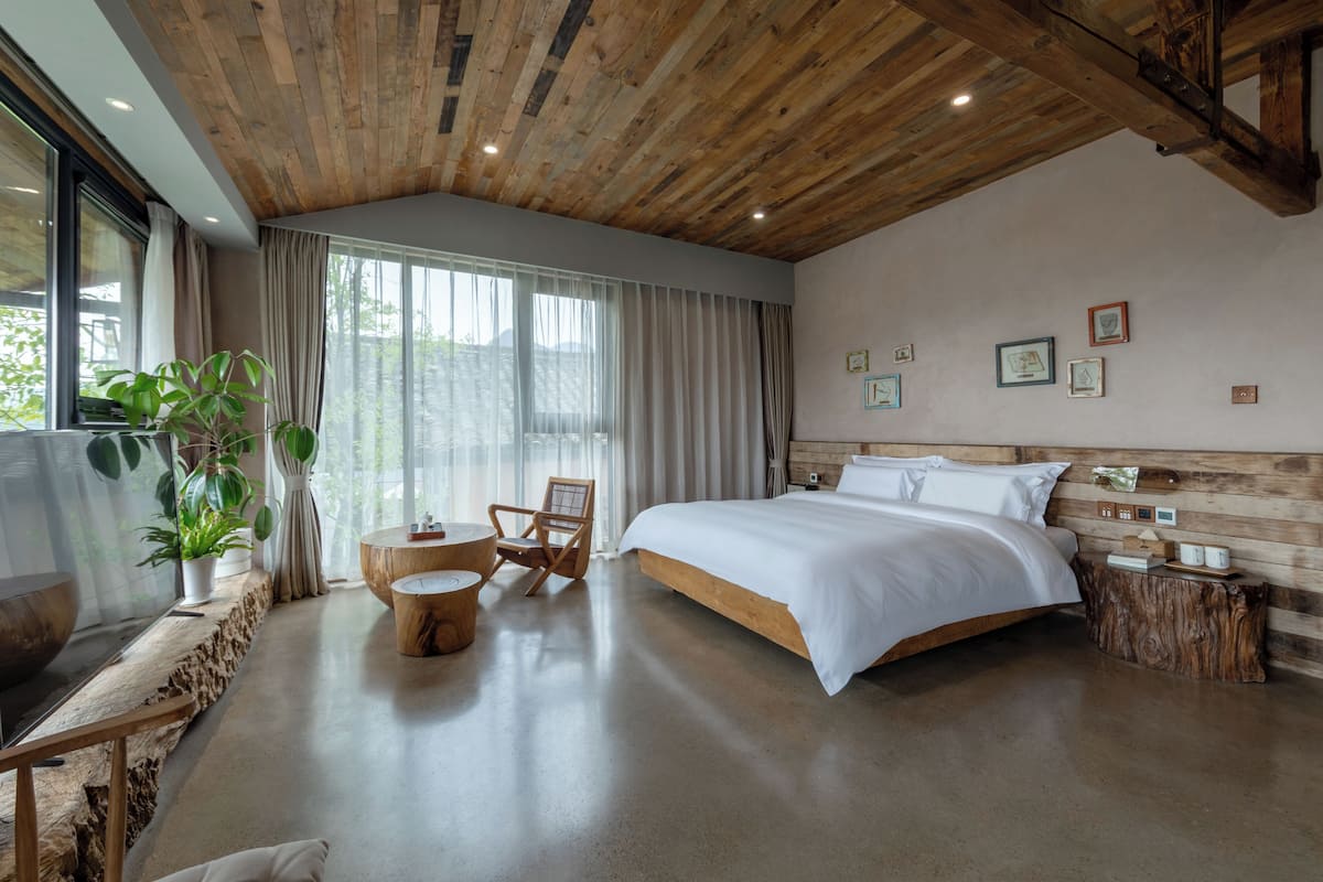 A bedroom with a wooden ceiling, a large bed, a TV, a plant, and a chair.