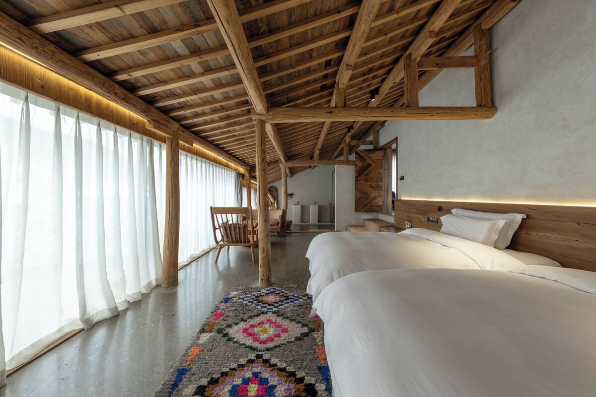 A bedroom with a wooden ceiling, a large bed, a chair, and a window with curtains.