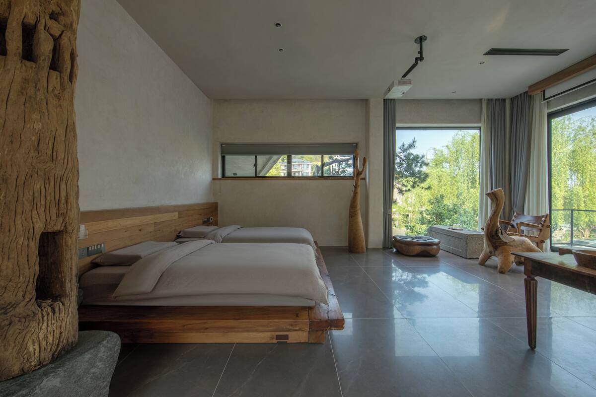A modern bedroom with a large bed, a wooden headboard, a window, and a view of trees.