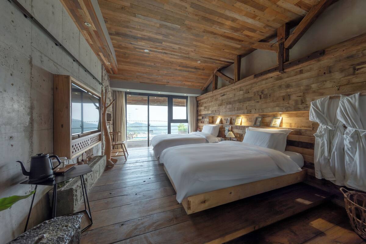 A wooden cabin-style bedroom with two beds, a small table with a kettle, and a view of the outdoors.