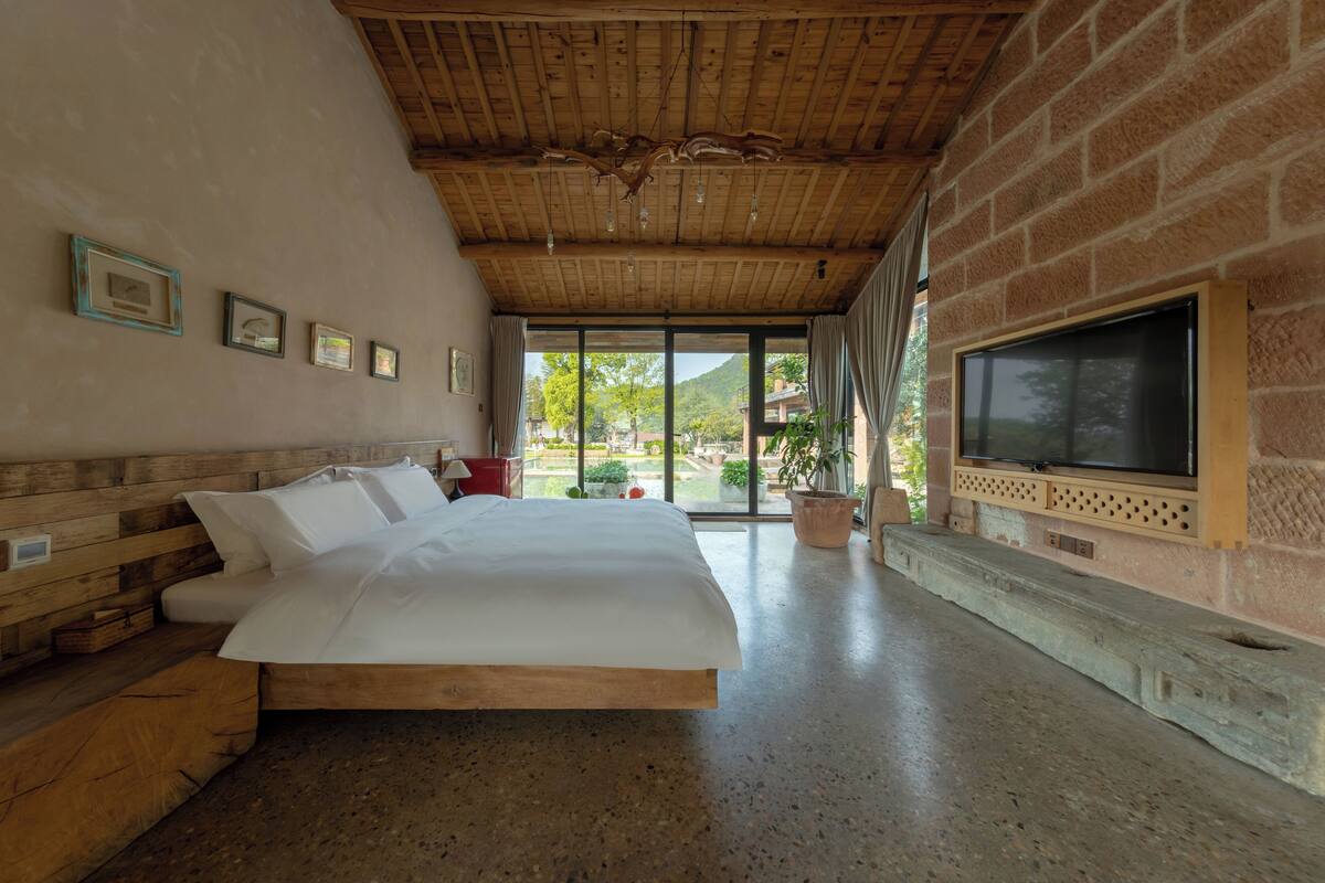 A bedroom with a wooden ceiling, a large bed, a TV on a stone fireplace, and a view of the outdoors.