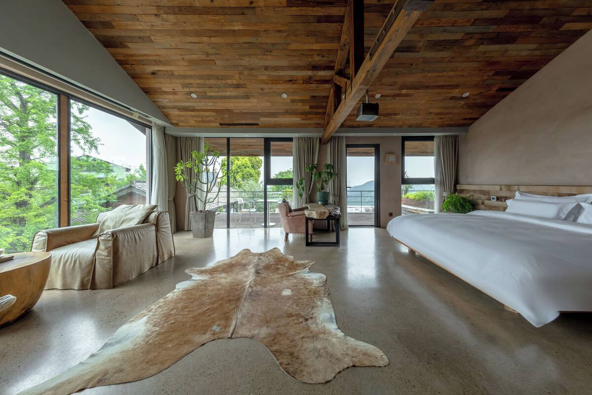 A spacious bedroom with a large bed, a leather sofa, a wooden ceiling, and a view of the outdoors.
