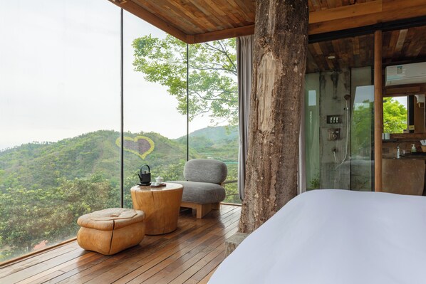 A bedroom with a large window, wooden flooring, a bed, a chair, and a small round table.