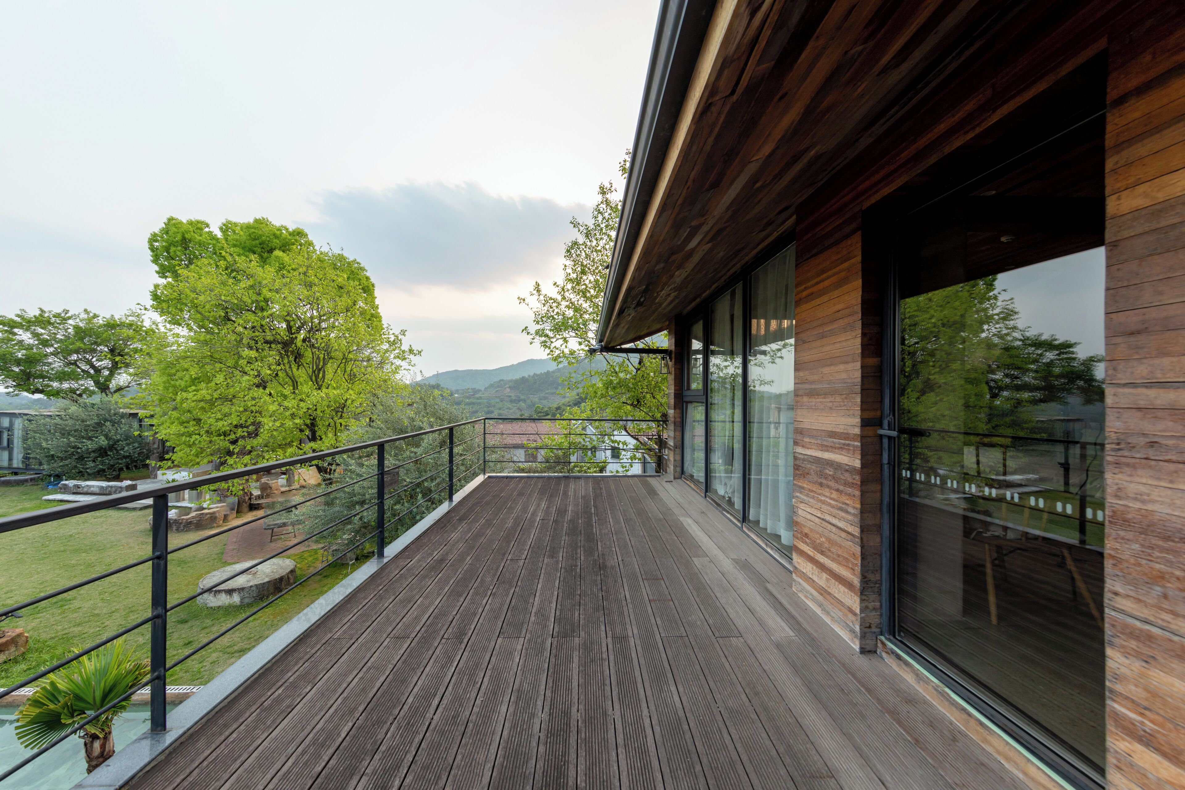 A wooden deck with a glass balustrade, a grassy area, and a view of trees and hills.