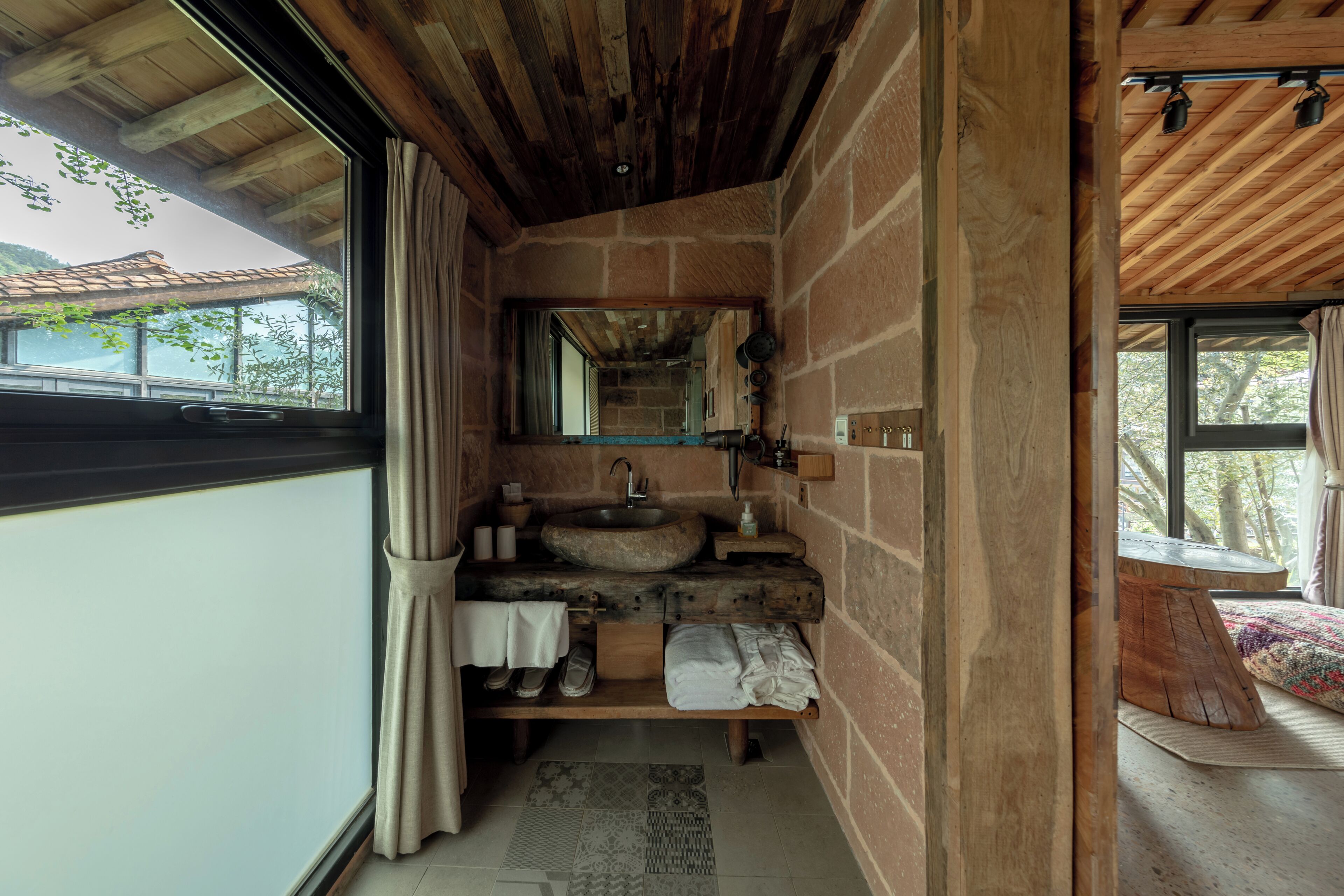 A rustic bathroom with a stone sink, wooden shelves, and a view of the outdoors.