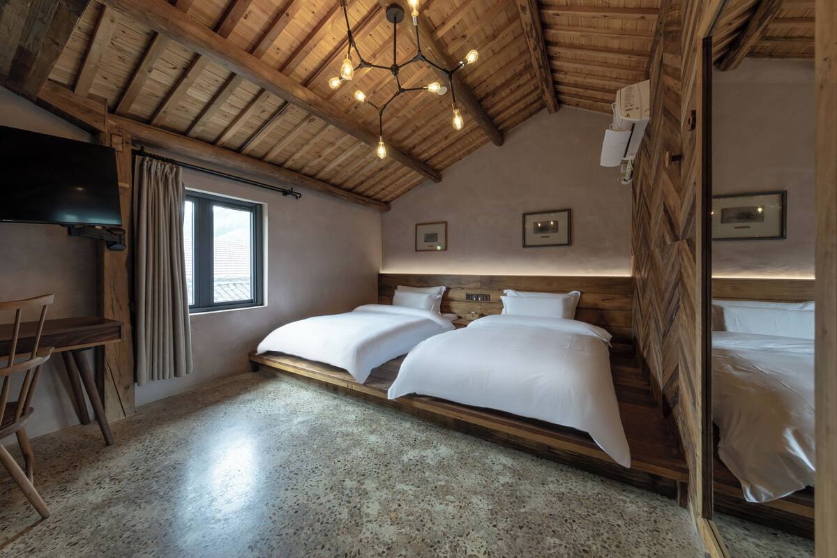 A bedroom with a wooden ceiling, two beds, a desk, and a window.