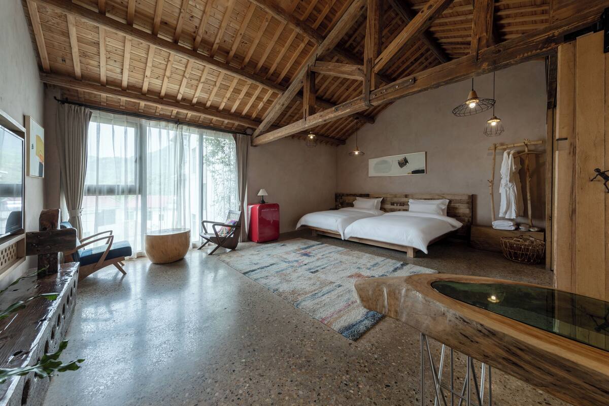 A bedroom with a large bed, wooden ceiling beams, a window with curtains, a small table, and a chair.