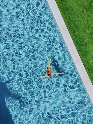 A person swimming in a rectangular pool with a clear blue surface, surrounded by a white pool edge and a green lawn. - Calarè Boutique Rooms (Castiadas)