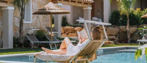 Una persona descansando en una silla de piscina, leyendo un libro, rodeada de un jardín cuidado y un edificio de piedra.