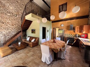 A spacious living area with a stone wall, a wooden staircase, a dining table with chairs, a kitchen with a refrigerator and stove, and a clock on the wall.