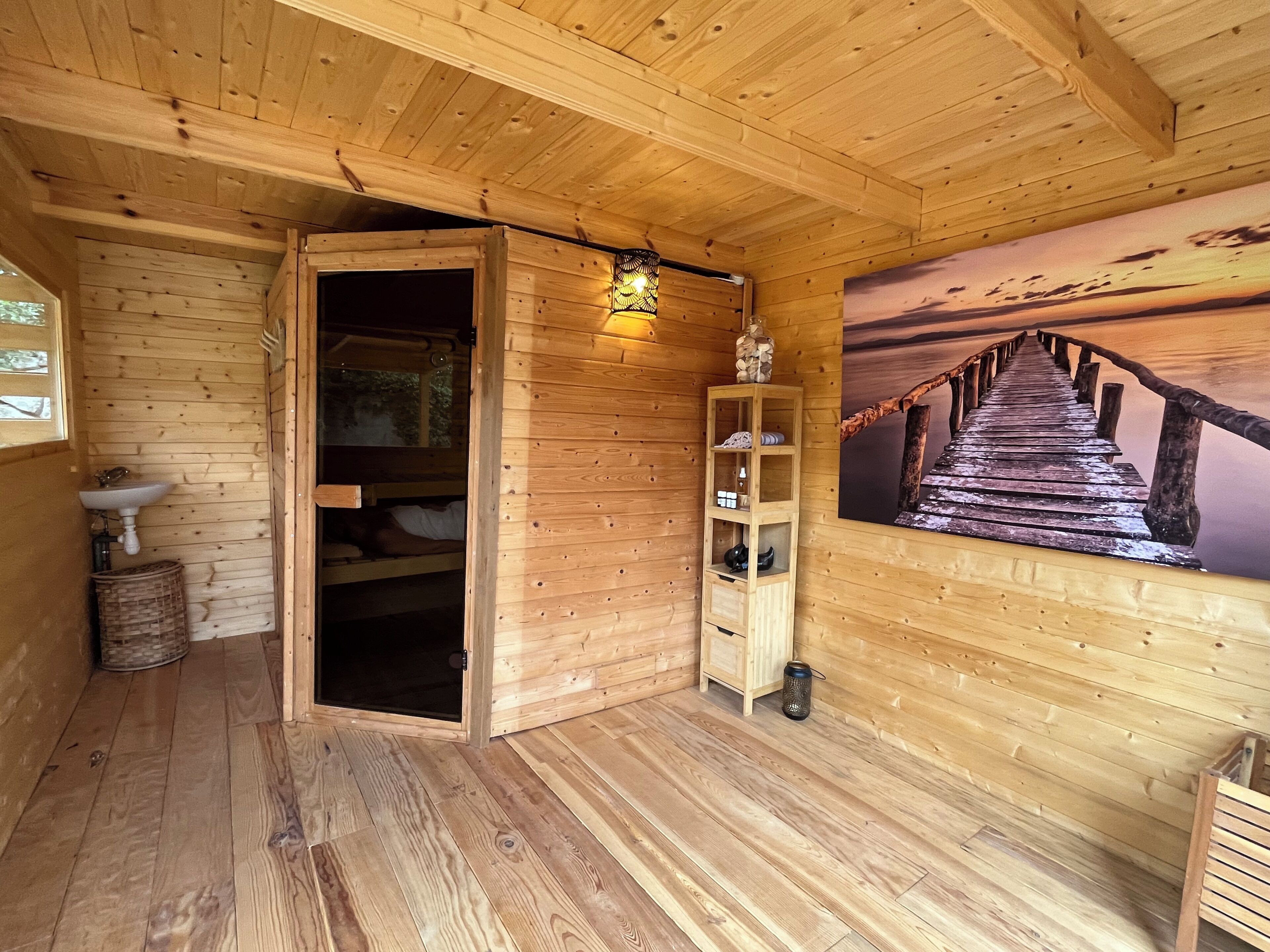 L’intérieur d’un chalet en bois, avec un banc en bois, une étagère ornée d’objets décoratifs, un tableau représentant un coucher de soleil et une fenêtre.