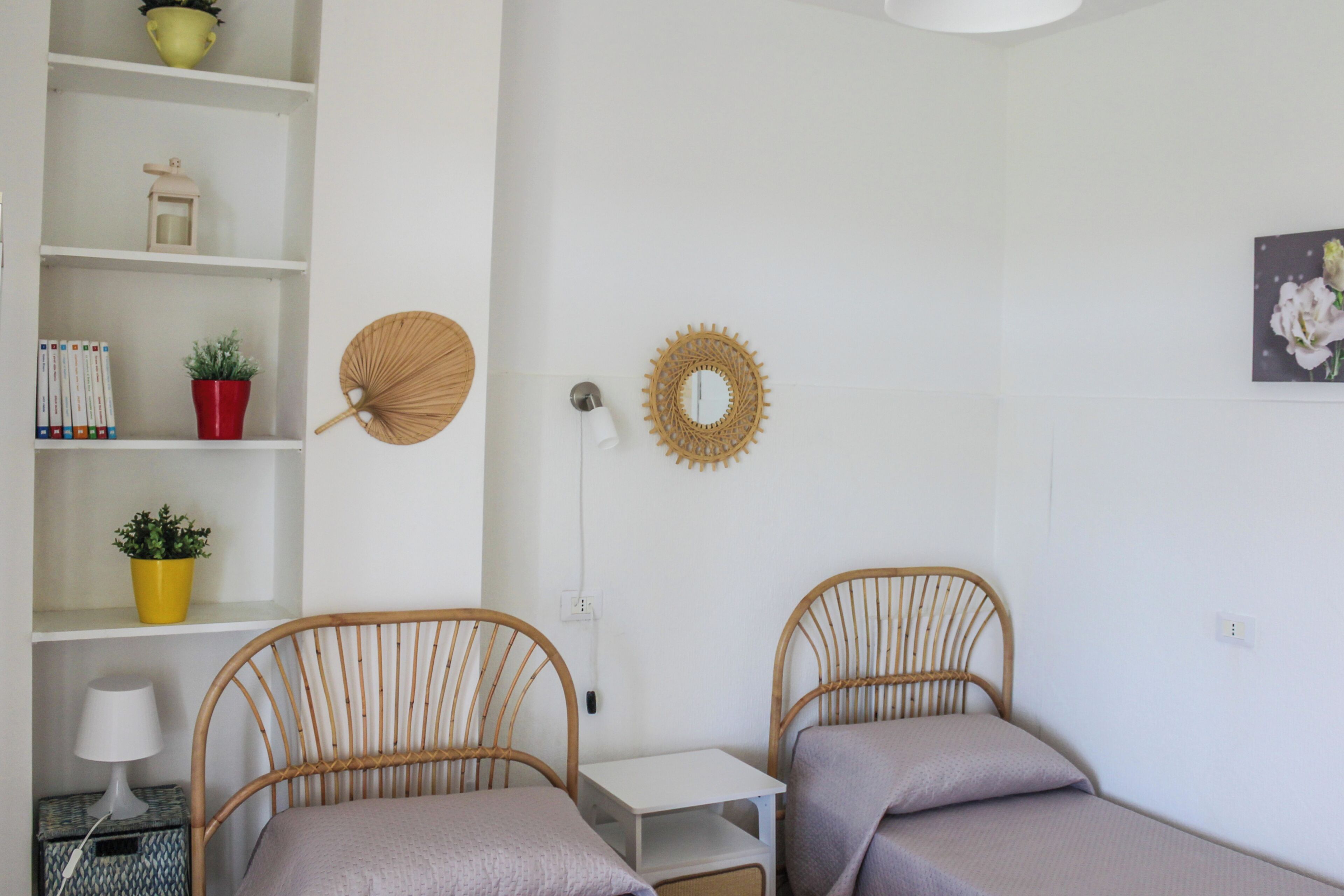 A room with two single beds, a small white table, a shelf with decorative items, and a wall-mounted mirror.