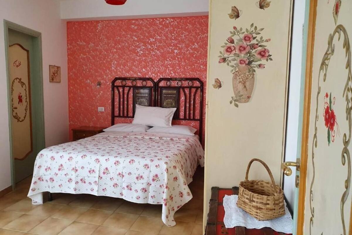 A bedroom with a bed, a red textured wall, a floral wall decoration, and a wicker basket on a table.