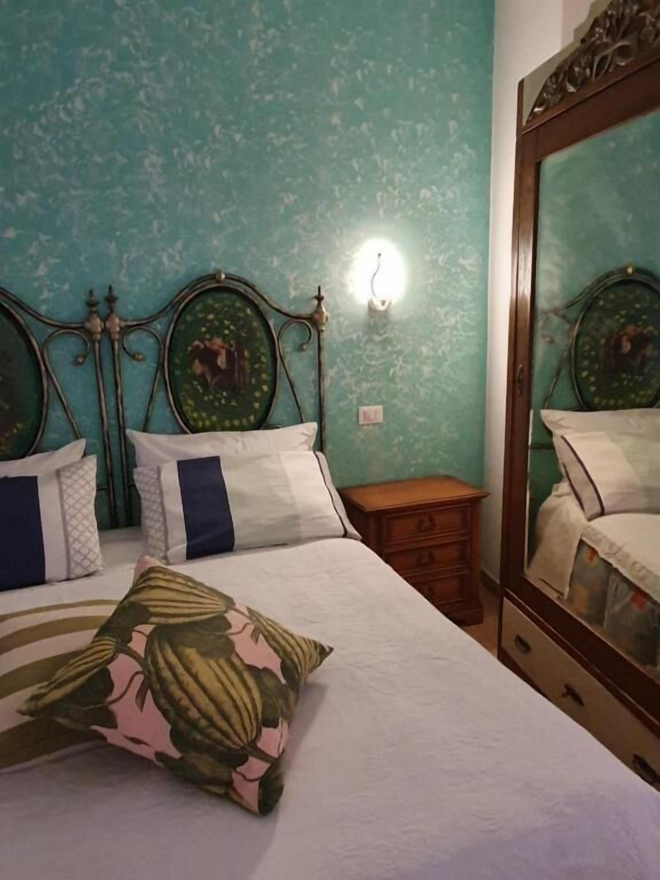 A bed with a floral-patterned pillow, a nightstand, a mirror, and a wall-mounted light fixture.