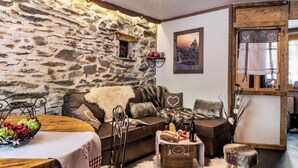 A cozy living room with a stone wall, a wooden table with fruit, a sofa with cushions, and a painting on the wall. - Your summer retreat in the heart of the mountains, services included (Aime-la-Plagne)