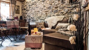 A cozy living room with a stone wall, a brown sofa, a dining table with checkered tablecloths, and a basket of fruit. - Your summer retreat in the heart of the mountains, services included (Aime-la-Plagne)