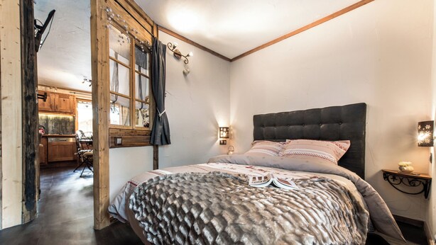 A bedroom with a large bed, wooden beams, a window with curtains, and a view of the kitchen. - Your summer retreat in the heart of the mountains, services included (Aime-la-Plagne)