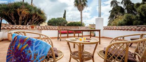 An outdoor patio with wicker furniture, a round table with food, and a view of palm trees and a chimney.