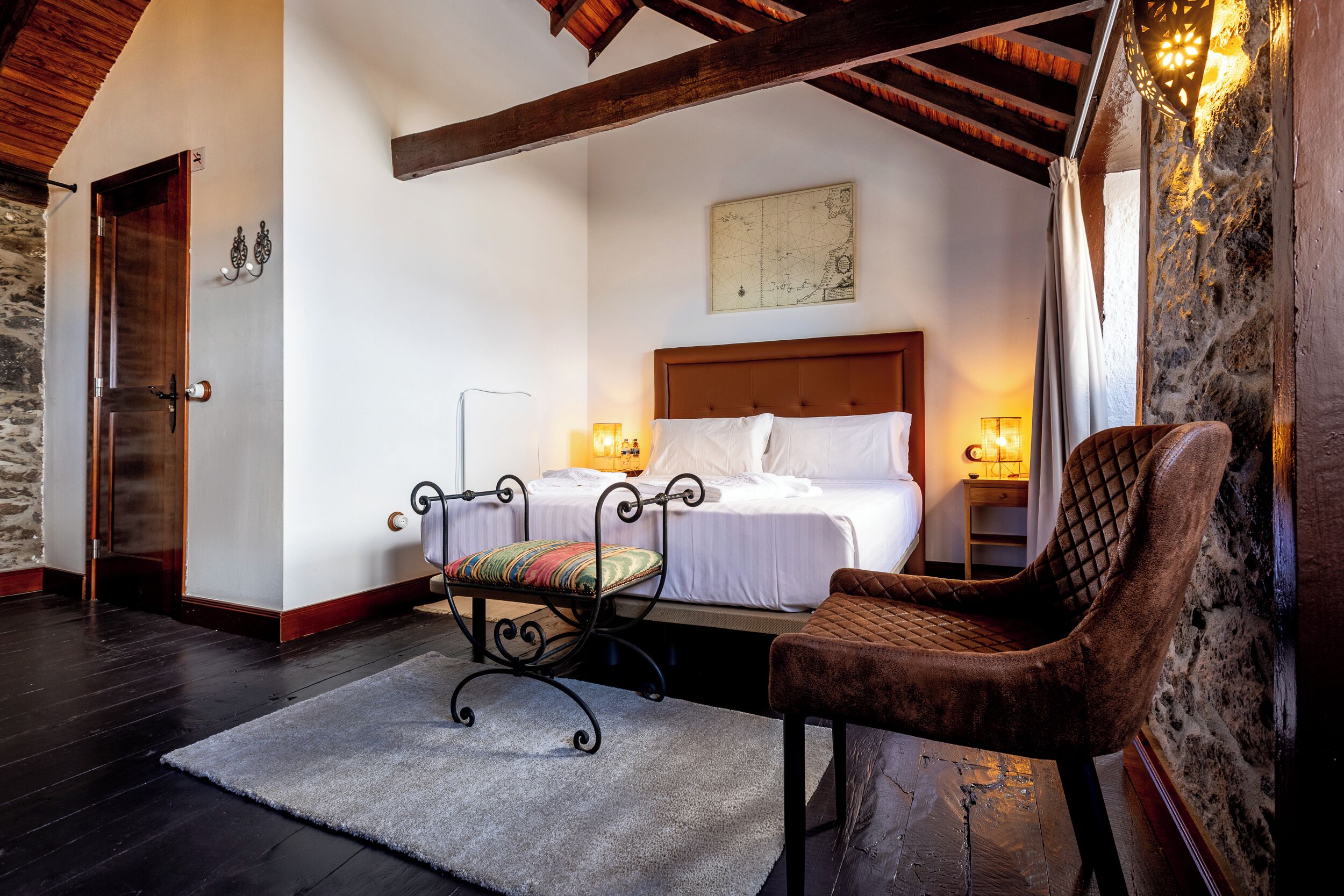 Een slaapkamer met een bed, twee bedlampen, een stoel, een vloerkleed en een stenen muur.
