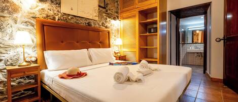 A hotel room with a stone wall, a wooden headboard, a bed with white linens, a nightstand with a lamp, and a wooden wardrobe.
