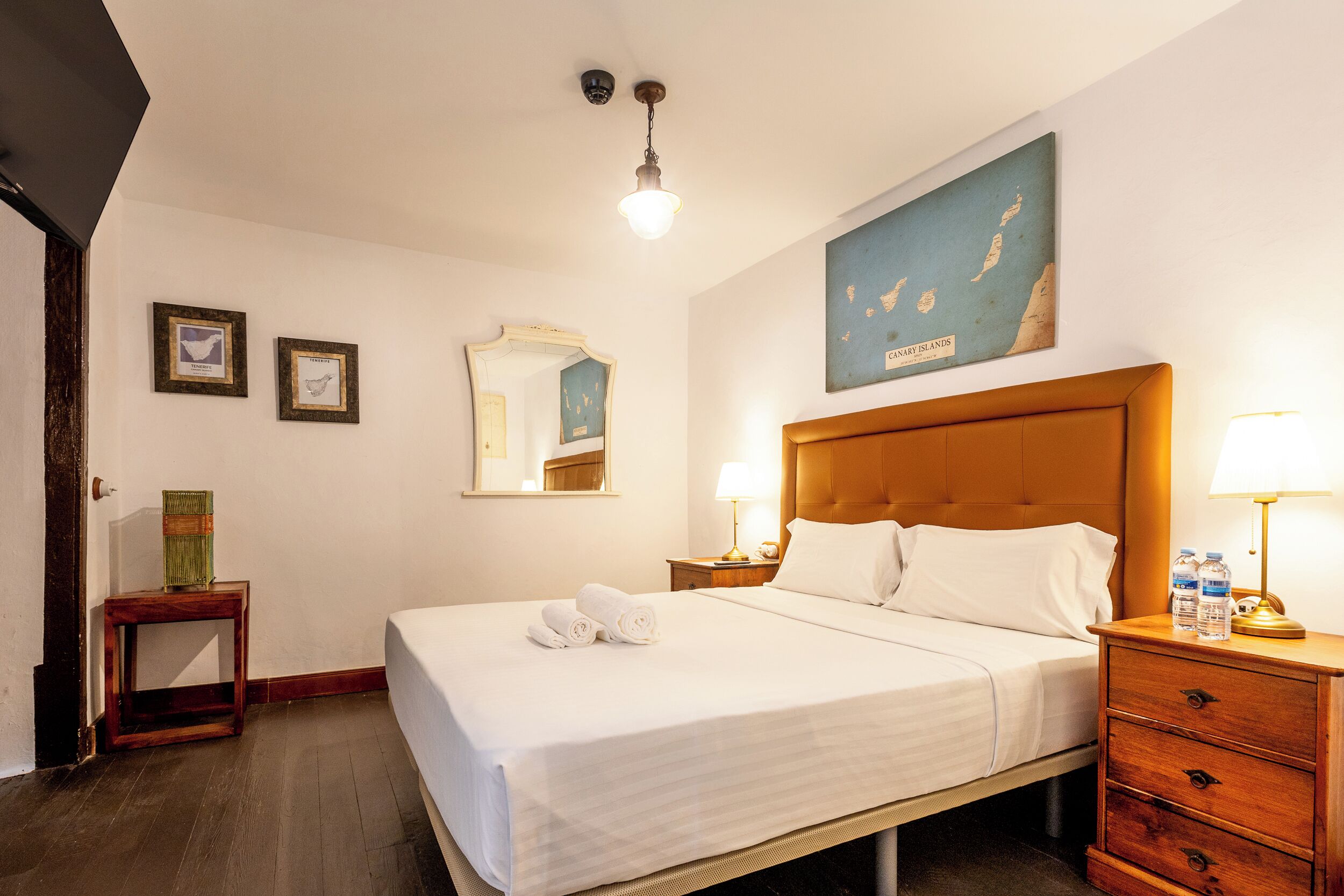 A hotel room with a bed, a nightstand with a lamp, a wooden dresser, a framed map on the wall, and a small table with a vase.