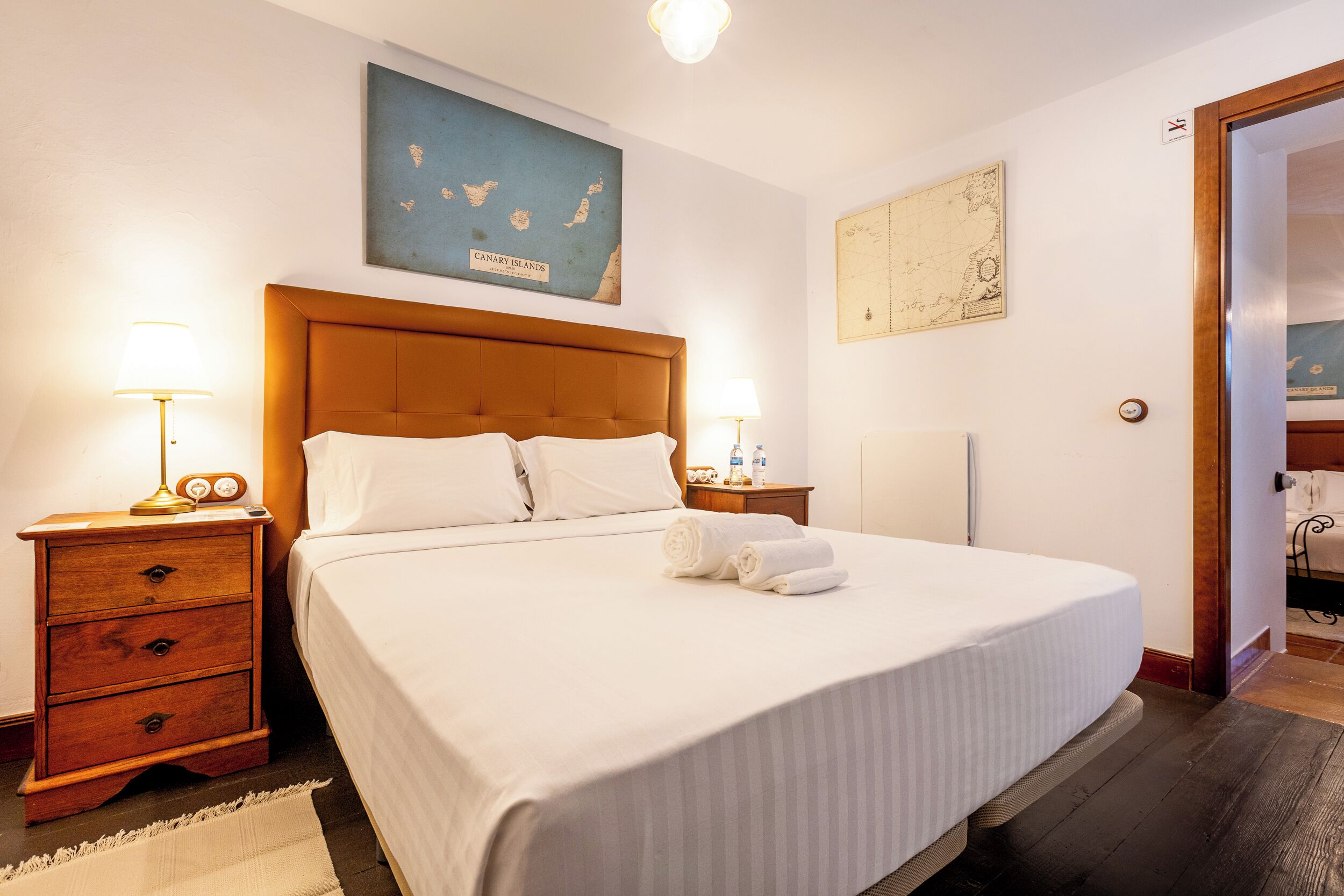 A neatly made bed with white linens, a wooden nightstand with a lamp, and a map on the wall.