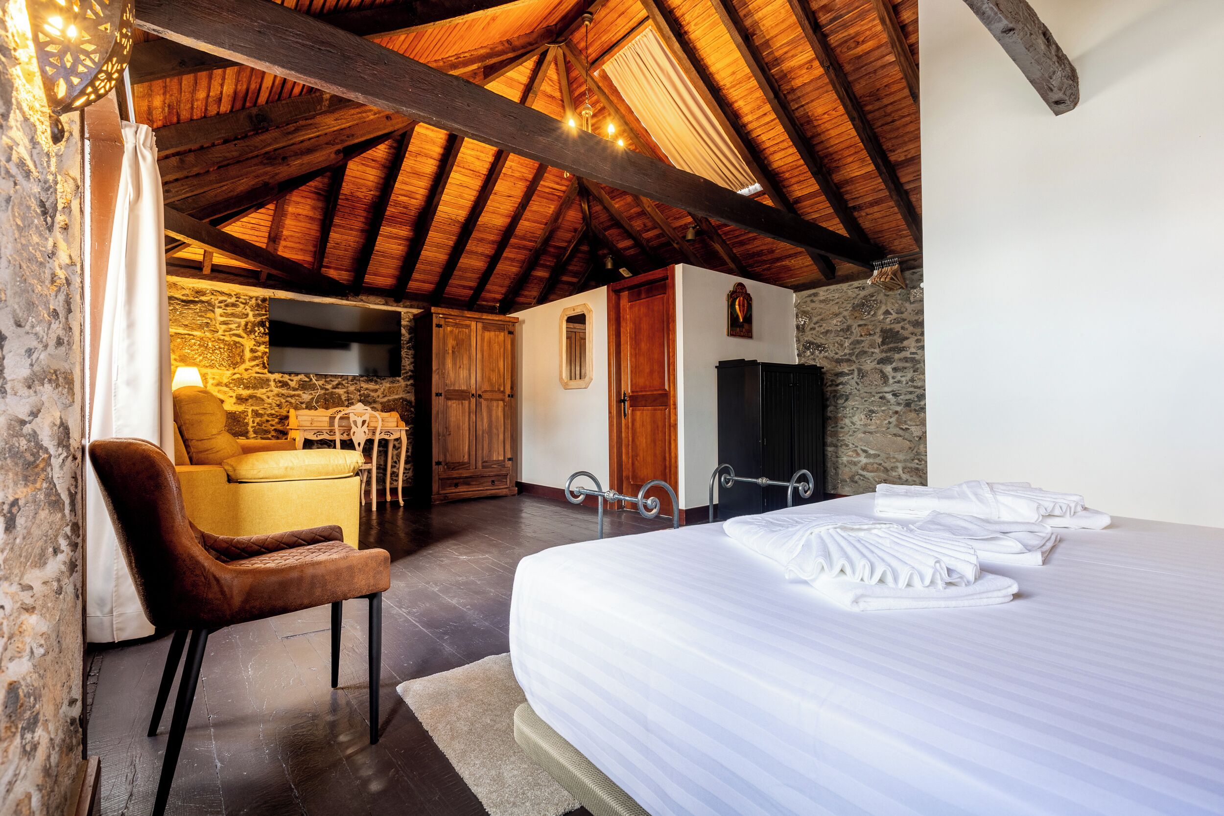 A bedroom with a large bed, a chair, a wooden ceiling, and a stone wall.