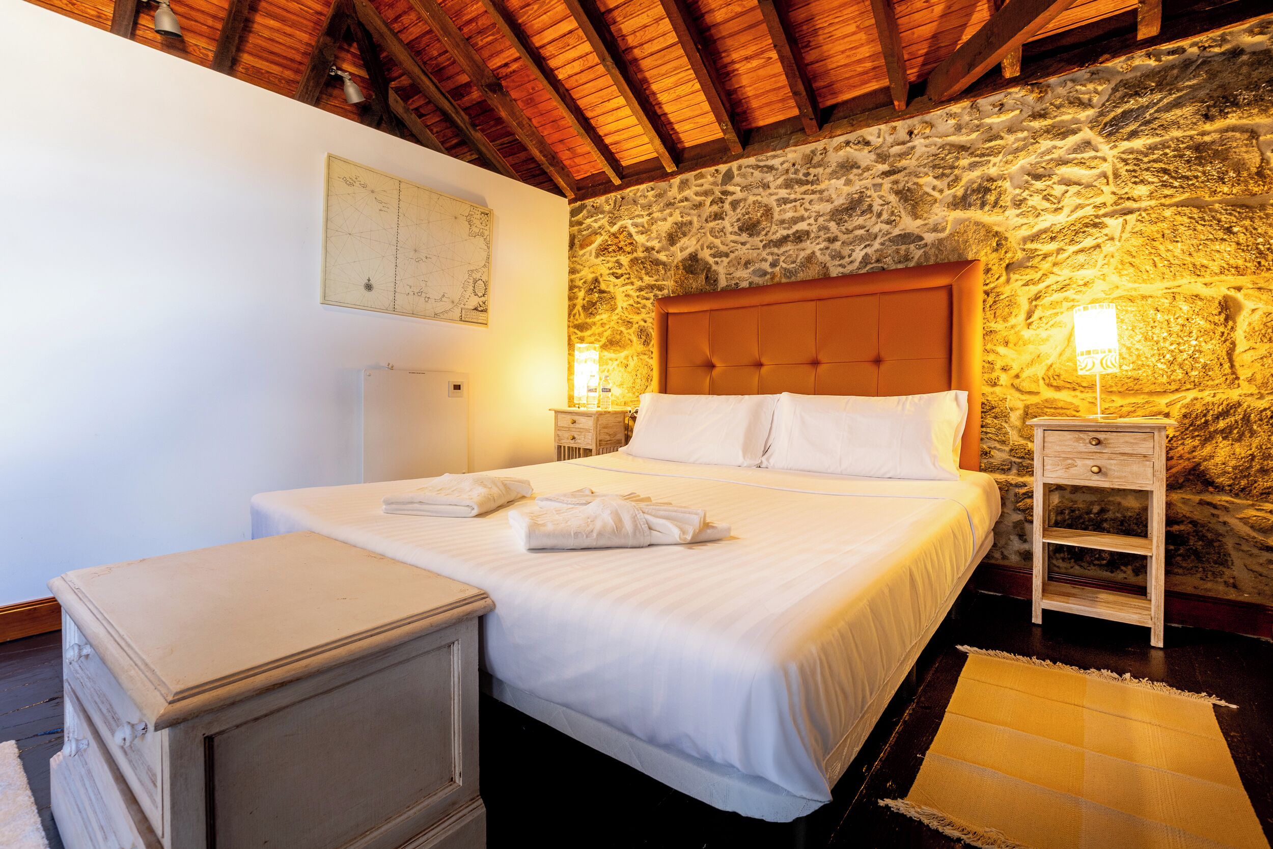 A bedroom with a stone wall, a wooden ceiling, a bed with white linens, a nightstand, and a map on the wall.