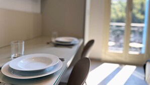 A dining table with plates and a glass, a chair, and a view of an outdoor area through a window. - Appartement 8A - MyResidenced'Azur (Beausoleil)