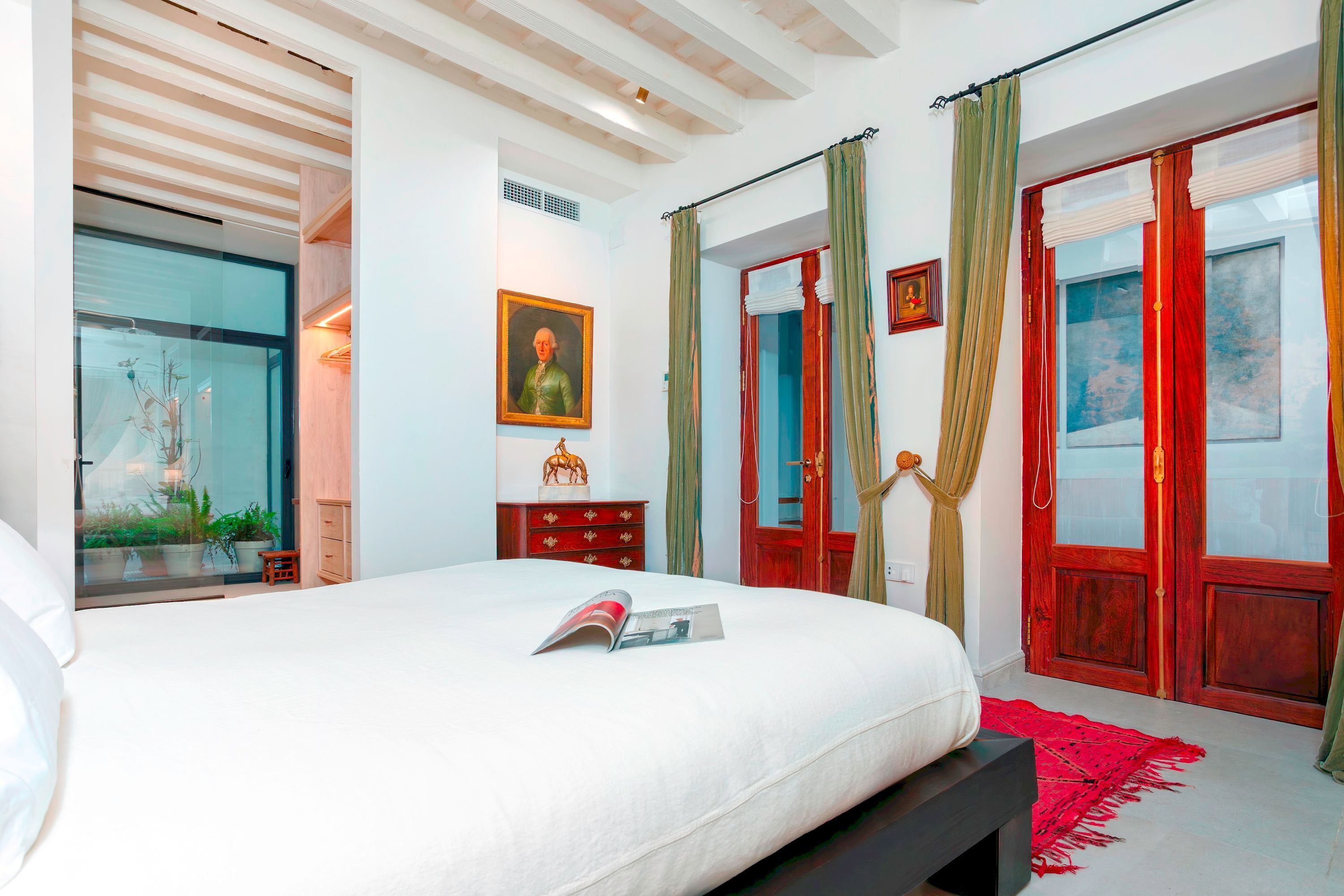 Un dormitorio con una cama, una puerta roja, una ventana con cortinas, un aparador de madera y un cuadro en la pared.