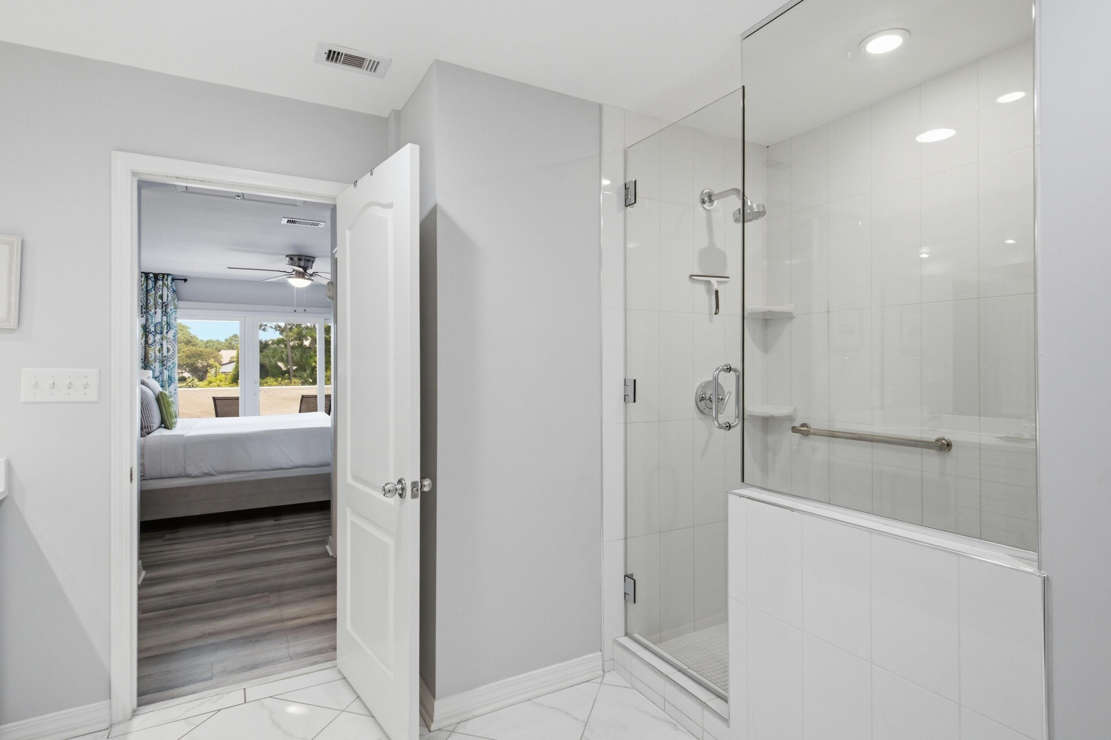 A modern bathroom with a glass shower enclosure, a visible door leading to a bedroom, and a ceiling fan.
