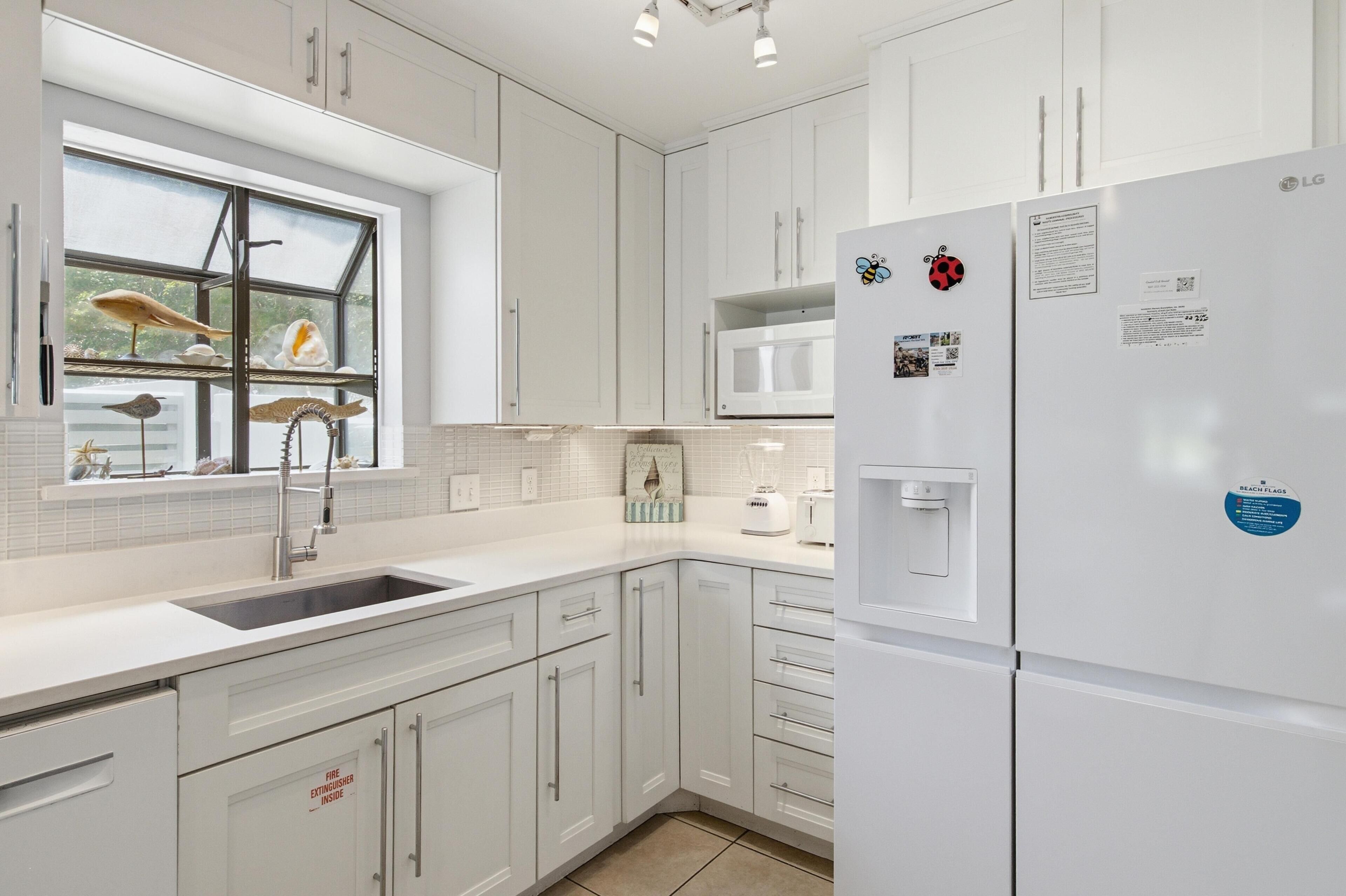 A modern kitchen with white cabinets, a built-in microwave, a refrigerator, and a window with decorative items.