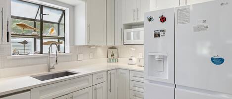 A modern kitchen with white cabinets, a built-in microwave, a refrigerator, and a window with decorative items.